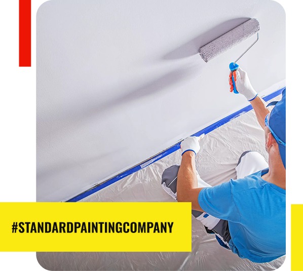 Person in blue shirt, white pants, gloves, and cap uses a paint roller to apply white paint to a wall. Floor is covered with plastic sheeting. Text reads #STANDARDPAINTINGCOMPANY—your trusted Painting Contractor—in a yellow banner.
