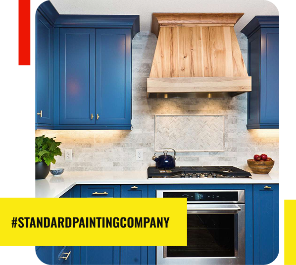 A modern kitchen with blue cabinets, a light wood range hood, white marble backsplash, and stainless steel oven. A potted plant and bowl of apples sit on the white countertop. #STANDARDPAINTINGCOMPANY—your trusted Painting Contractor—is on a yellow banner.