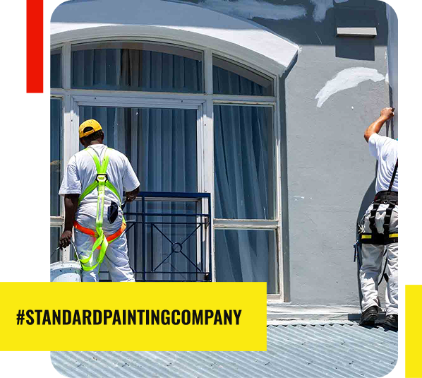 Two painters in white workwear are painting the exterior of a building with large arched windows. One holds a bucket, while the other uses a brush. A yellow banner reads #STANDARDPAINTINGCOMPANY, your trusted painting contractor in Park Ridge.