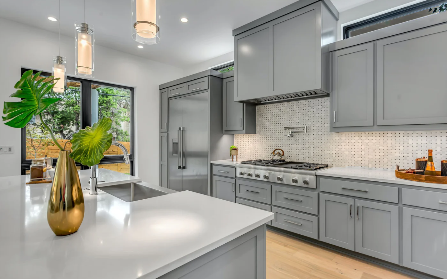 Modern kitchen with gray cabinets, stainless steel appliances, and a white countertop island with a sink. Enhanced by pendant lights and a gold vase with green leaves, this space shines—expertly finished by Standard Painting Company for a flawless look.