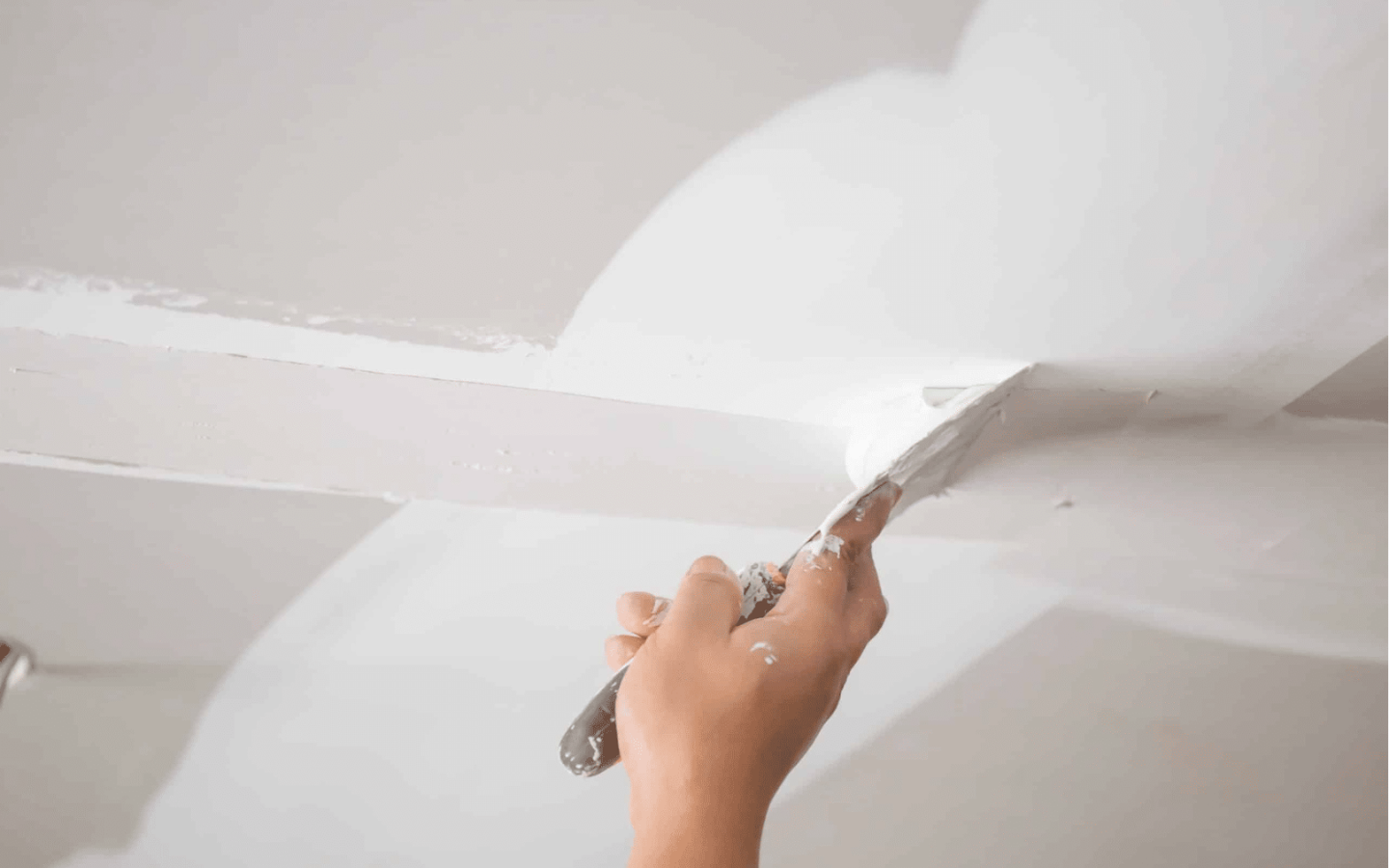 A hand uses a putty knife to apply white joint compound to a ceiling, expertly smoothing over seams between drywall panels—a task often handled by a professional Painting Contractor like Standard Painting Company.