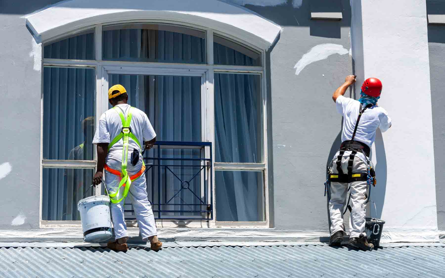 Two workers in safety gear from Standard Painting Company paint the exterior of a building near a large arched window. One holds a bucket, while the other uses a paintbrush on the wall. Both stand on a corrugated metal roof.